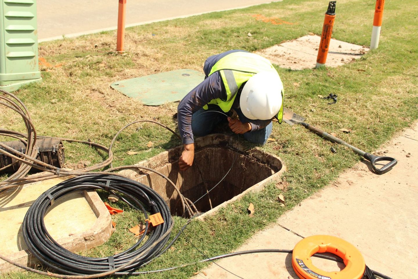 A One World Communications technician in a hard hat and high visibility vest working an open handhole